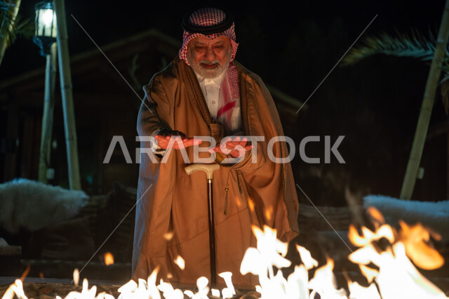 جلسة وكشتة بر في منطقة طبيعية بالمملكة العربية السعودية، إشعال موقد الحطب للتدفئة، الأجواء الشتوية الباردة، رجل عربي خليجي سعودي يرتدي الزي التقليدي والشماغ والفروة في الجو البارد يقف أمام شعلة النار، قضاء وقت ممتع في الإجازة