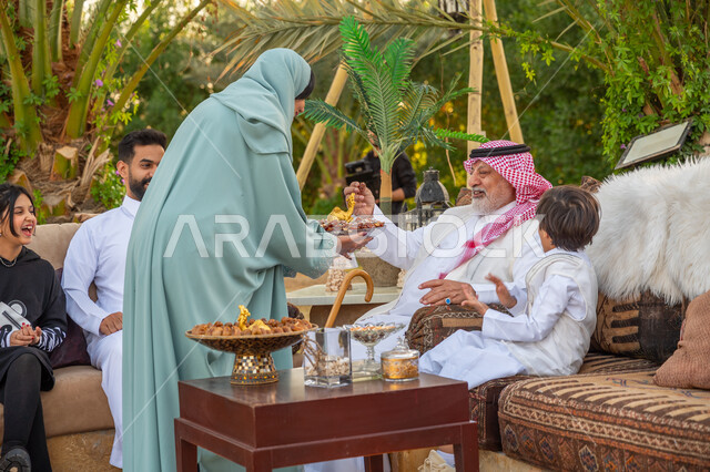 الأجواء الأسرية السعيدة, تبادل الأحاديث والأخبار بإيماءات السرور, تقوية وتعزيز الرابط الأسري, امرأة عربية خليجية سعودية محجبة ترتدي عباءة زرقاء تقوم بتقديم الرطب لرجل كبير السن, جلسة عائلية بالهواء الطلق