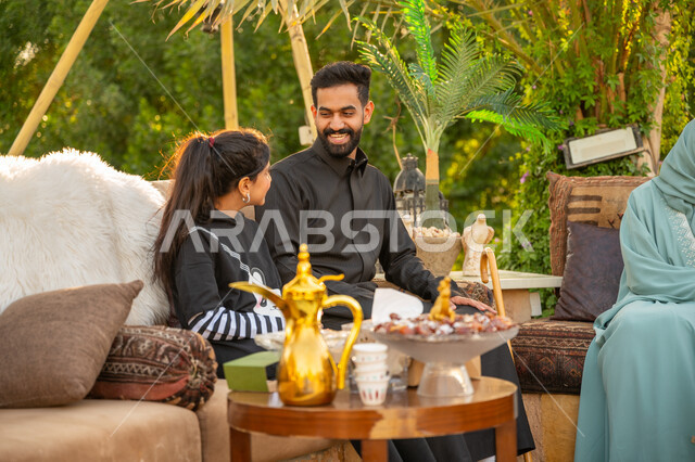 جلسة عائلية بالهواء الطلق, الأجواء الأسرية السعيدة, تبادل الأحاديث والأخبار بإيماءات السرور, تقوية وتعزيز الرابط الأسري, الاستمتاع بالمشروب الساخن, أب عربي خليجي سعودي يرتدي الثوب التقليدي يجلس مع ابنته في الحديقة في النهار، قضاء الوقت الممتع