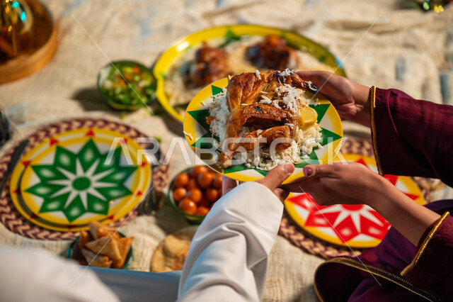 الأجواء الرمضانية الروحانية، اجتماع عائلي على سفرة الإفطار في شهر رمضان المبارك، صورة مقربة ليدين إمرأة عربية خليجية سعودية تقوم بسكب صحن من إرز المندي مع الدجاج لزوجها، تقديم الطعام للاسرة