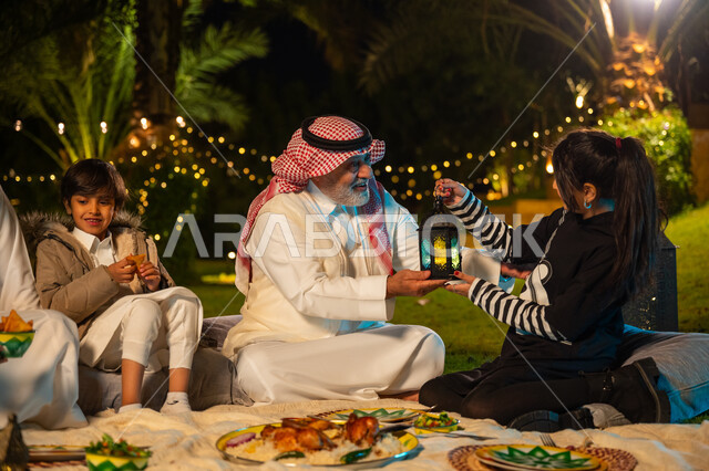 Ramadan decorations, enjoying breakfast outdoors with family, elderly Saudi Arabian Gulf man wearing traditional thobe and shemagh giving his granddaughter a decorated metal lantern, concept of family bonding, family gathering at the dining table, happy family Ramadan atmosphere