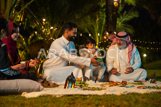 Happy family Ramadan atmosphere, Ramadan decorations, enjoying breakfast outdoors with family, family gathering at the table, concept of family bonding, Saudi Arabian Gulf man wearing traditional dress offering dates to his father