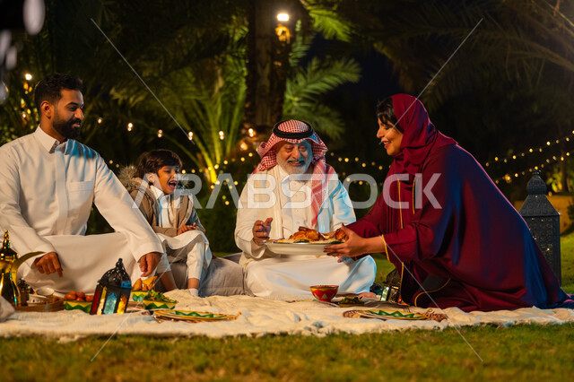 مفهوم الترابط الأسري, اجتماع الأسرة على السفرة, عائلة عربية خليجية سعودية يتناولون الطعام في الحديقة, امرأة ترتدي عباءة حمراء تقوم بتقديم طبق الأكل, الأجواء الرمضانية العائلية السعيدة, زينة شهر رمضان المبارك, الاستمتاع بتناول الإفطار بالهواء الطلق مع الأهل