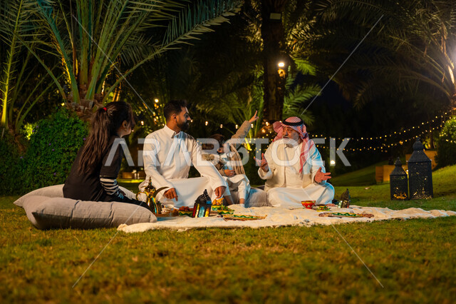 الاستمتاع بتناول الإفطار بالهواء الطلق, مفهوم الترابط الأسري, اجتماع الأسرة على السفرة, الأجواء الرمضانية العائلية السعيدة, زينة وديكور شهر رمضان المبارك, رجل كبير السن عربي خليجي سعودي يرتدي الثوب التقليدي والشماغ يجلس بجانب ابنه ويتبادل الأحاديث والأخبار