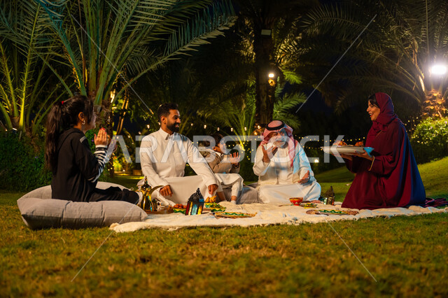 زينة شهر رمضان المبارك, الاستمتاع بتناول الإفطار بالهواء الطلق مع الأهل, مفهوم الترابط الأسري, اجتماع الأسرة على السفرة, عائلة عربية خليجية سعودية يتناولون الطعام في الحديقة, امرأة ترتدي عباءة حمراء تقوم بتقديم طبق الأكل, الأجواء الرمضانية العائلية السعيدة