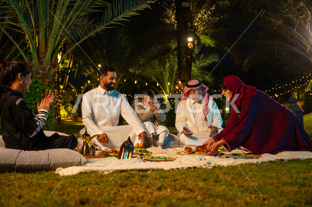 الأجواء الرمضانية العائلية السعيدة, زينة شهر رمضان المبارك, الاستمتاع بتناول الإفطار بالهواء الطلق مع الأهل, مفهوم الترابط الأسري, اجتماع الأسرة على السفرة, عائلة عربية خليجية سعودية يتناولون الطعام في الحديقة, امرأة ترتدي عباءة حمراء تقوم بتقديم طبق الأكل