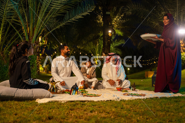 Ramadan decorations, enjoying breakfast outdoors with family, concept of family bonding, family gathering at the table, Saudi Arabian Gulf family eating in the garden, woman wearing red abaya serving food, happy family Ramadan atmosphere