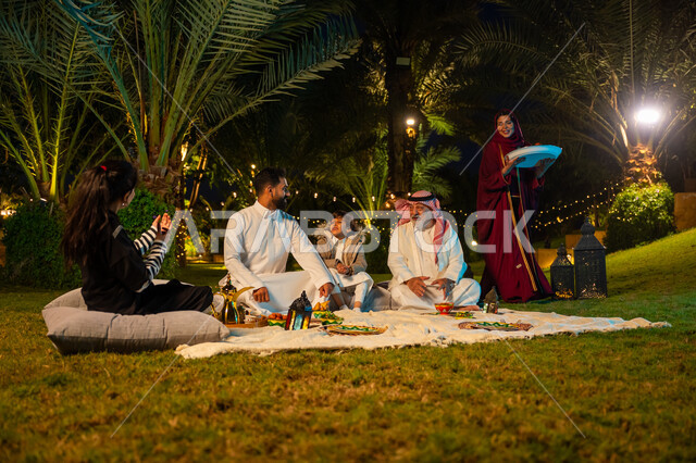 Happy family Ramadan atmosphere, Ramadan decorations, enjoying breakfast outdoors with family, concept of family bonding, family gathering at the table, Saudi Arabian Gulf family eating in the garden, woman wearing red abaya serving food