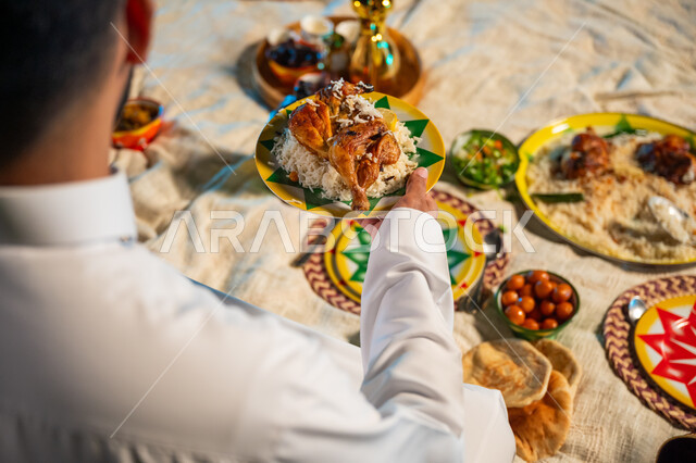 سفرة رمضانية متنوعة بالمأكولات والمقبلات الشهية، صورة مقربة من الخلف لرجل عربي خليجي سعودي يرتدي الثوب التقليدي يجلس على مائدة الافطار ويحمل بيده صحن من أرز المندي مع الدجاج، الأجواء الرمضانية الروحانية