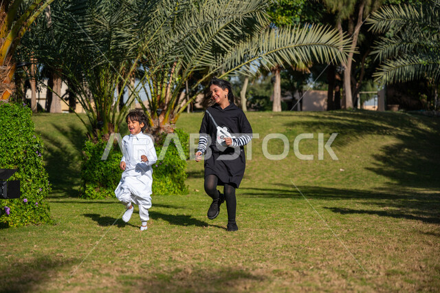 اللعب واللهو والمرح, قضاء الوقت الممتع مع الأخوة, الاستمتاع بالتنزه في الهواء الطلق, حديقة ومنتزه ترفيهي عائلي, فتاة عربية خليجية سعودية ترتدي الزي الكاجوال تركض في إحدى الحدائق مع أخيها الصغير في النهار