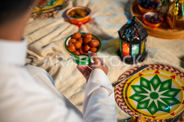 فانوس معدني مضيء مع دلة من القهوة والفناجين، حلى رمضاني سعودي شهير، صورة مقربة من الخلف لرجل عربي يحمل بيده صحن مليء بحلوى اللقيمات الشهية، حسن الضيافة والاستقبال في شهر رمضان المبارك، الأجواء الرمضانية الروحانية