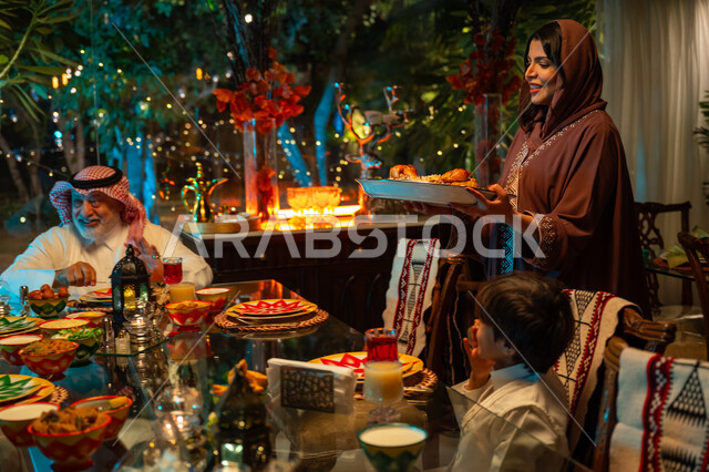 تبادل الأحاديث بين أفراد الأسرة، أداء فريضة الصيام في شهر رمضان المبارك، سفرة مليئة بأصناف من المأكولات الشرقية الشهية، أجواء عائلية دينية رمضانية سعيدة، عائلة عربية خليجية سعودية مجتمعة حول مائدة الطعام، تقديم الطبق الرئيسي للافطار، زينة المناسبات الإسلامية