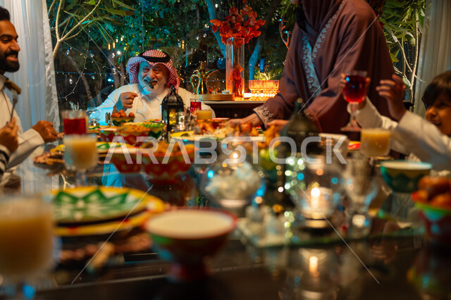 سفرة مليئة بأصناف من المأكولات الشرقية الشهية، أجواء عائلية دينية رمضانية سعيدة، عائلة عربية خليجية سعودية مجتمعة حول مائدة الطعام، أداء فريضة الصيام في شهر رمضان المبارك، تبادل الأحاديث بين أفراد الأسرة، تقديم الطبق الرئيسي للافطار، زينة المناسبات الإسلامية