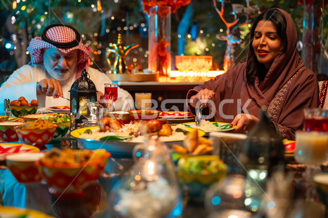 أجواء عائلية دينية رمضانية سعيدة، ديكور وزينة المناسبات الإسلامية، تبادل الأحاديث بين أفراد الأسرة، أداء فريضة الصيام في شهر رمضان المبارك، سفرة مليئة بأصناف من المأكولات الشرقية الشهية، عائلة عربية خليجية سعودية مجتمعة حول مائدة الطعام