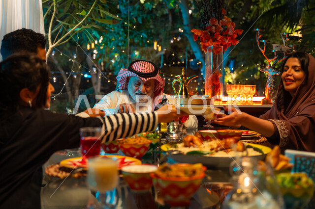 سفرة مليئة بأصناف من المأكولات الشرقية الشهية، أجواء عائلية دينية رمضانية سعيدة، عائلة عربية خليجية سعودية مجتمعة حول مائدة الطعام، أداء فريضة الصيام في شهر رمضان المبارك، تبادل الأحاديث بين أفراد الأسرة، زينة المناسبات الإسلامية