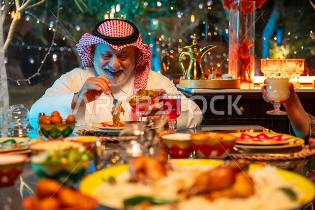 Spending quality time with family at a table full of delicious food and drinks, gestures of happiness and joy at the family gathering, an elderly Saudi Arabian Gulf man wearing traditional thobe and shemagh eating Ramadan breakfast at sunset, a spiritual Ramadan family gathering, decorations and decor for the holy month of Ramadan