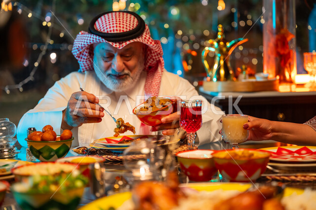 ايماءات السعادة والسرور بلمة الأهل، رجل مسن عربي خليجي سعودي يرتدي الثوب التقليدي والشماغ يقوم بتناول الافطار الرمضاني عند أذان المغرب, قضاء وقت ممتع مع العائلة على مائدة مليئة بالأطعمة والمشروبات الشهية, جلسة عائلية رمضانية روحانية، زينة وديكور شهر رمضان المبارك