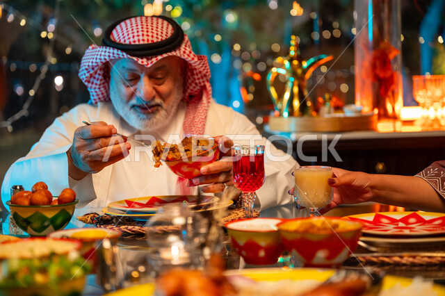 تعابير السعادة والسرور بلمة الأهل، رجل مسن عربي خليجي سعودي يرتدي الثوب التقليدي والشماغ يقوم بتناول الافطار الرمضاني عند أذان المغرب, قضاء وقت ممتع مع العائلة على مائدة مليئة بالأطعمة والمشروبات الشهية, جلسة عائلية رمضانية روحانية، زينة وديكور شهر رمضان المبارك
