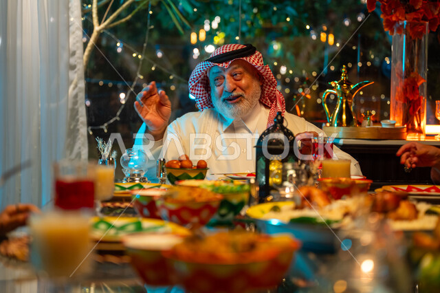 تعابير السعادة والسرور بلمة الأهل، تناول الافطار الرمضاني عند أذان المغرب, رجل مسن عربي خليجي سعودي يرتدي الثوب التقليدي والشماغ يقضي وقت ممتع مع اسرته على مائدة مليئة بالأطعمة والمشروبات الشهية, جلسة عائلية رمضانية روحانية، زينة وديكور شهر رمضان المبارك