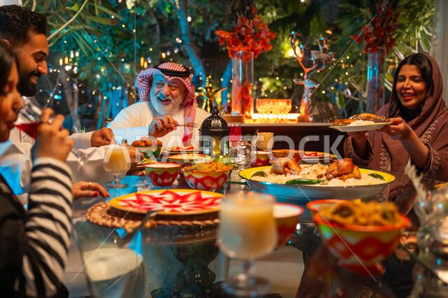 ايماءات السعادة والسرور بلمة الأهل، ديكور وزينة شهر رمضان المبارك، جلسة عائلية رمضانية روحانية، مائدة مليئة بالأطعمة والمشروبات الشهية, تناول الافطار الرمضاني عند أذان المغرب, امرأة عربية خليجية سعودية تجلس مع عائلتها وتقوم بسكب صحن من الأرز مع الدجاج المشوي