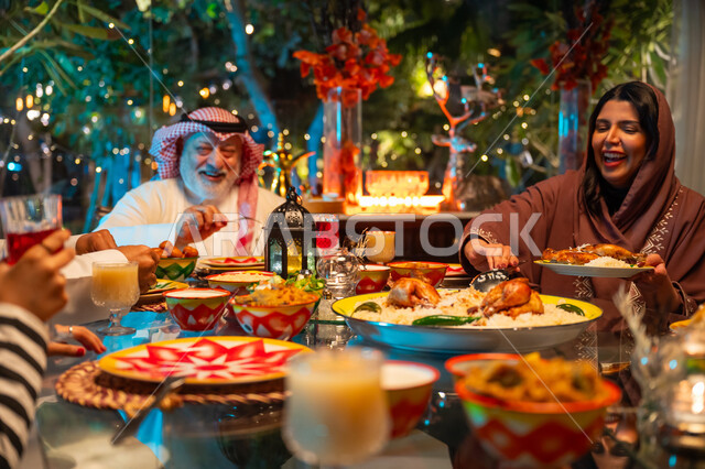 تعابير السعادة والسرور بلمة الأهل، ديكور وزينة شهر رمضان المبارك، جلسة عائلية رمضانية روحانية، مائدة مليئة بالأطعمة والمشروبات الشهية, تناول الافطار الرمضاني عند أذان المغرب, امرأة عربية خليجية سعودية تجلس مع عائلتها وتقوم بسكب صحن من الأرز مع الدجاج المشوي