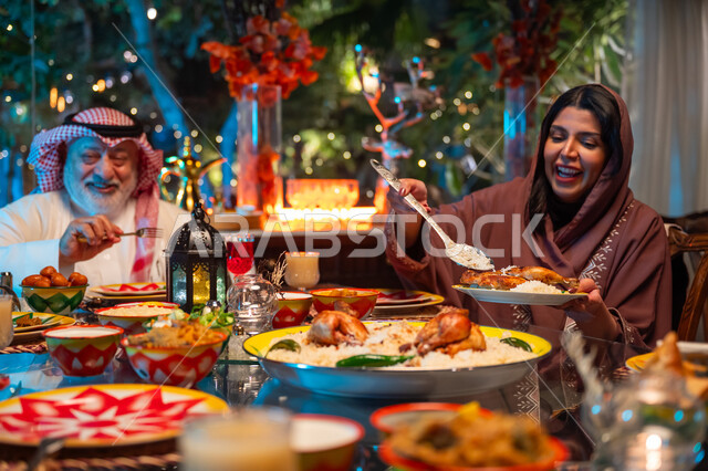 ديكور وزينة شهر رمضان المبارك، جلسة عائلية رمضانية روحانية، مائدة مليئة بالأطعمة والمشروبات الشهية, تناول الافطار الرمضاني عند أذان المغرب, تعابير السعادة والسرور بلمة الأهل، امرأة عربية خليجية سعودية تجلس مع عائلتها وتقوم بسكب صحن من الأرز مع الدجاج المشوي