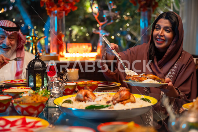 زينة وديكور شهر رمضان المبارك، جلسة عائلية رمضانية روحانية، مائدة مليئة بالأطعمة والمشروبات الشهية, تناول الافطار الرمضاني عند أذان المغرب, تعابير السعادة والسرور بلمة الأهل، امرأة عربية خليجية سعودية تقوم بسكب صحن من الأرز مع الدجاج المشوي