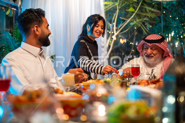 سفرة مليئة بالأطعمة والمشروبات الشهية, قضاء وقت ممتع مع الأسرة، جلسة عائلية رمضانية روحانية، زينة وديكور شهر رمضان المبارك، تعابير السعادة والسرور بلمة الأهل، عائلة عربية خليجية سعودية تجلس على طاولة الطعام لتناول الافطار الرمضاني عند أذان المغرب