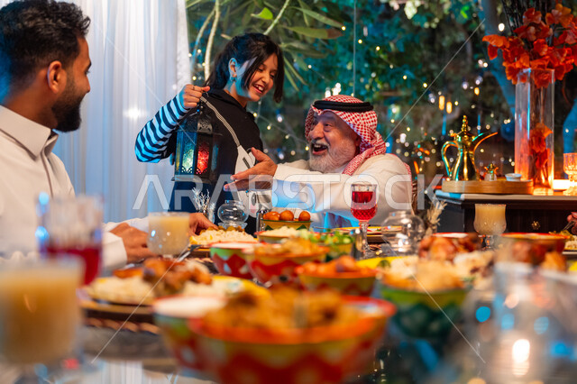 تعابير السعادة والسرور بلمة الأهل، مائدة مليئة بالأطعمة والمشروبات الشهية, الاستمتاع بقضاء الوقت مع الأسرة، جلسة عائلية رمضانية روحانية، ديكور وزينة شهر رمضان المبارك، عائلة عربية خليجية سعودية تجلس على طاولة الطعام لتناول الافطار الرمضاني عند أذان المغرب