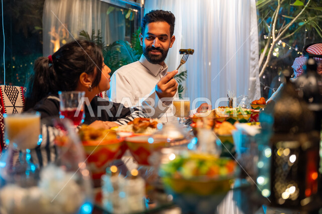 مائدة مليئة بالأطعمة والمشروبات الشهية, الاستمتاع بقضاء الوقت مع الأسرة، جلسة عائلية رمضانية روحانية، ديكور وزينة شهر رمضان المبارك، ايماءات السعادة والسرور بلمة الأهل، عائلة عربية خليجية سعودية تجلس على طاولة الطعام لتناول الافطار الرمضاني عند أذان المغرب