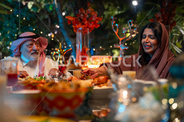 زينة وديكور شهر رمضان المبارك، جلسة عائلية رمضانية روحانية، قضاء وقت ممتع مع الأسرة، مائدة مليئة بالأطعمة والمشروبات الشهية, ايماءات السعادة والسرور بلمة الأهل، عائلة عربية خليجية سعودية تجلس على طاولة الطعام لتناول الافطار الرمضاني عند أذان المغرب
