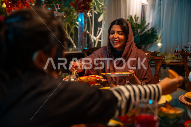 سفرة مليئة بالأطعمة والمشروبات الشهية, قضاء وقت ممتع مع الأسرة، جلسة عائلية رمضانية روحانية، زينة وديكور شهر رمضان المبارك، تعابير السعادة والسرور بلمة الأهل، عائلة عربية خليجية سعودية تجلس على طاولة الطعام لتناول الافطار الرمضاني عند أذان المغرب