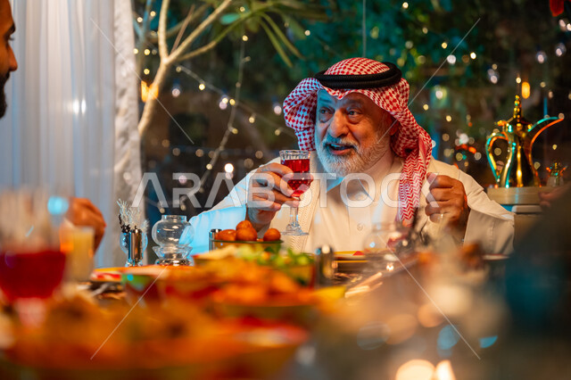 Eating Ramadan breakfast at sunset, gestures of happiness and joy at the family gathering, an elderly Saudi Arabian Gulf man wearing the traditional thobe and shemagh spending quality time with his family at a table full of delicious food and drinks, a spiritual Ramadan family gathering, decorations and decor for the holy month of Ramadan