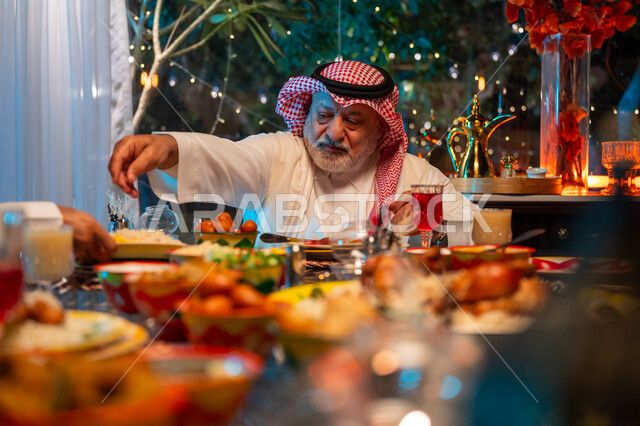ايماءات السعادة والسرور بلمة الأهل، تناول الافطار الرمضاني عند أذان المغرب, رجل مسن عربي خليجي سعودي يرتدي الثوب التقليدي والشماغ يقضي وقت ممتع مع اسرته على مائدة مليئة بالأطعمة والمشروبات الشهية, جلسة عائلية رمضانية روحانية، زينة وديكور شهر رمضان المبارك