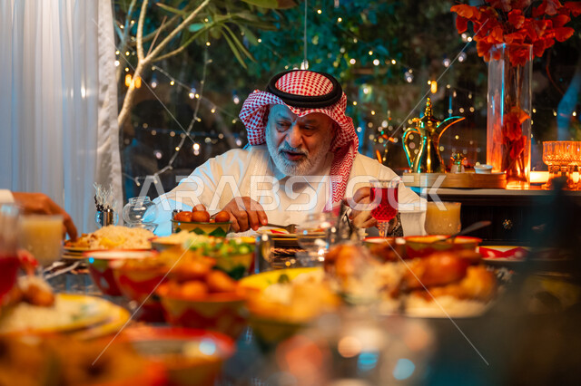 Expressions of happiness and joy at the family gathering, eating Ramadan breakfast at sunset, an elderly Saudi Arabian Gulf man wearing the traditional thobe and shemagh spending quality time with his family at a table full of delicious food and drinks, a spiritual Ramadan family gathering, decorations and decor for the holy month of Ramadan
