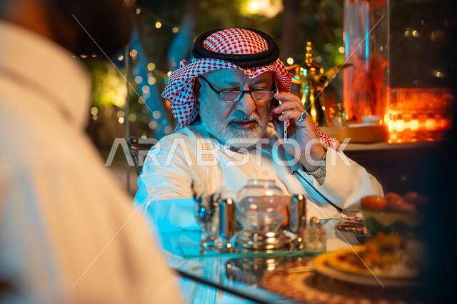 التواصل مع الاصدقاء للتهنئة بقدوم شهر الخير، جلسة عائلية رمضانية روحانية، تناول الافطار الرمضاني عند أذان المغرب، رجل مسن عربي خليجي سعودي يرتدي الثوب التقليدي والشماغ يجلس على مائدة الفطور ويقوم باجراء مكالمة عبر الهاتف المحمول، زينة وديكور شهر رمضان المبارك