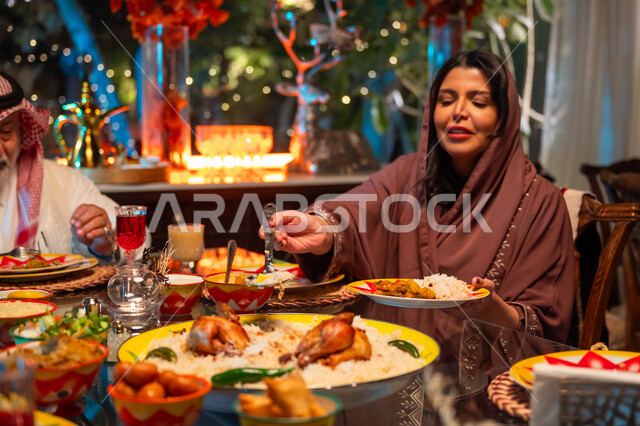 مائدة مليئة بالأطعمة والمشروبات الشهية, تناول الافطار الرمضاني عند أذان المغرب, تعابير السعادة والسرور بلمة الأهل، ديكور وزينة شهر رمضان المبارك، جلسة عائلية رمضانية روحانية، امرأة عربية خليجية سعودية تجلس مع عائلتها وتقوم بسكب صحن من الأرز مع الدجاج المشوي