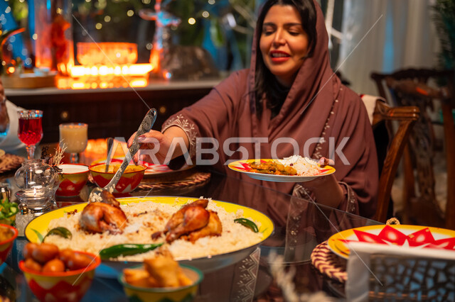 تناول الافطار الرمضاني عند أذان المغرب, تعابير السعادة والسرور بلمة الأهل، ديكور وزينة شهر رمضان المبارك، جلسة عائلية رمضانية روحانية، مائدة مليئة بالأطعمة والمشروبات الشهية, امرأة عربية خليجية سعودية تجلس مع عائلتها وتقوم بسكب صحن من الأرز مع الدجاج المشوي