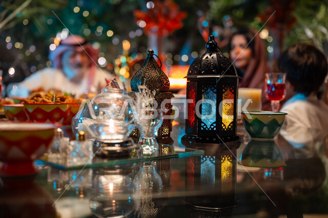 تبادل الأحاديث بين أفراد الأسرة، أجواء عائلية دينية رمضانية سعيدة، فوانيس معدنية مزخرفة ملونة على سفرة مليئة بأصناف من المأكولات الشرقية الشهية، زينة المناسبات الإسلامية، أداء فريضة الصيام في شهر رمضان المبارك، عائلة عربية خليجية سعودية مجتمعة حول مائدة الطعام