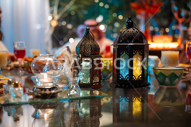 أجواء عائلية دينية رمضانية سعيدة، فوانيس معدنية مزخرفة ملونة على سفرة مليئة بأصناف من المأكولات الشرقية الشهية، زينة المناسبات الإسلامية، أداء فريضة الصيام في شهر رمضان المبارك، تبادل الأحاديث بين أفراد الأسرة، عائلة عربية خليجية سعودية مجتمعة حول مائدة الطعام