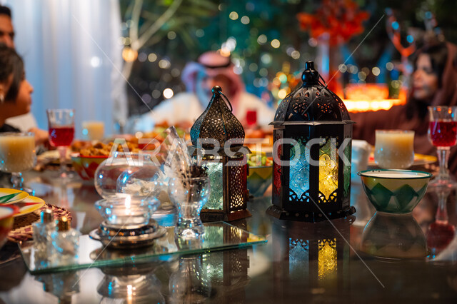 فوانيس معدنية مزخرفة ملونة على سفرة مليئة بأصناف من المأكولات الشرقية الشهية، زينة المناسبات الإسلامية، أداء فريضة الصيام في شهر رمضان المبارك، أجواء عائلية دينية رمضانية سعيدة، تبادل الأحاديث بين أفراد الأسرة، عائلة عربية خليجية سعودية مجتمعة حول مائدة الطعام