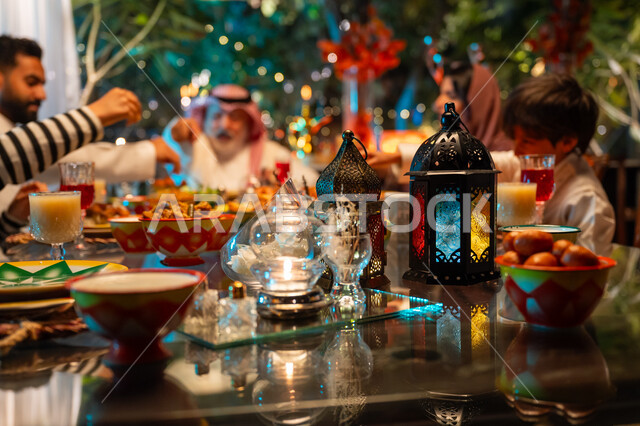 Performing the fasting duty in the holy month of Ramadan, a happy religious Ramadan family atmosphere, exchanging conversations between family members, colorful decorated metal lanterns on a table filled with delicious oriental foods, a Saudi Arabian Gulf family gathered around the dining table, Islamic occasion decorations