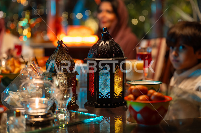 فوانيس معدنية مزخرفة ملونة على سفرة مليئة بأصناف من المأكولات الشرقية الشهية، تبادل الأحاديث بين أفراد الأسرة، أجواء عائلية دينية رمضانية سعيدة، عائلة عربية خليجية سعودية مجتمعة حول مائدة الطعام، أداء فريضة الصيام في شهر رمضان المبارك، زينة المناسبات الإسلامية