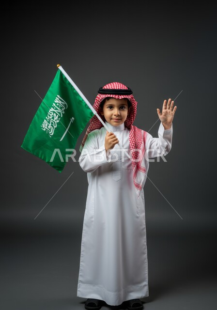 يوم العلم 11 مارس، مفهوم حب الوطن والانتماء اليه، ذكرى اليوم الوطني السعودي 23 سبتمبر، رفع اليد بتعابير القاء التحية والسلام، بورتريه لولد عربي خليجي سعودي يرتدي الثوب التقليدي والشماغ يحمل بيده علم المملكة، الجسم بالطول الكامل، خلفية رمادية