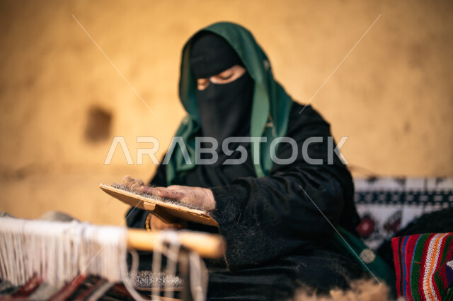 الشغف في ممارسة مهنة الغزل و النسج, حرفة يدوية تاريخيه, يوم بدينا 22 فبراير، المحافظة على التراث الشعبي السعودي, ذكرى تأسيس الدولة السعودية الأولى 1727م, صورة مقربة لامرأة عربيه خليجية سعودية ترتدي البرقع و العباءه السوداء تجلس في بيت من الطين تقوم بلف خيوط الصوف