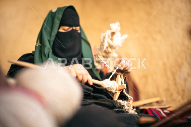 الشغف في ممارسة مهنة الغزل و النسج, حرفة يدوية تاريخيه, يوم بدينا 22 فبراير، المحافظة على التراث الشعبي السعودي, ذكرى تأسيس الدولة السعودية الأولى 1727م, صورة مقربة لامرأة عربيه خليجية سعودية ترتدي البرقع و العباءه السوداء تجلس في بيت من الطين تقوم بلف خيوط الصوف