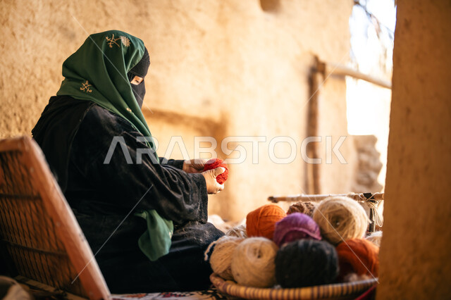 يوم بدينا 22 فبراير، ذكرى تأسيس الدولة السعودية الأولى 1727م, حرفة يدوية تاريخيه, الشغف في ممارسة مهنة الغزل و النسج, المحافظة على التراث الشعبي السعودي, صورة من الجانب لامرأة عربيه خليجية سعودية ترتدي البرقع و العباءه السوداء تجلس في بيت من الطين تقوم بلف خيوط الصوف