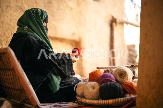 ذكرى تأسيس الدولة السعودية الأولى 1727م, حرفة يدوية تاريخيه, يوم بدينا 22 فبراير، الشغف في ممارسة مهنة الغزل و النسج, المحافظة على التراث الشعبي السعودي, صورة من الجانب لامرأة عربيه خليجية سعودية ترتدي البرقع و العباءه السوداء تجلس في بيت من الطين تقوم بلف خيوط الصوف