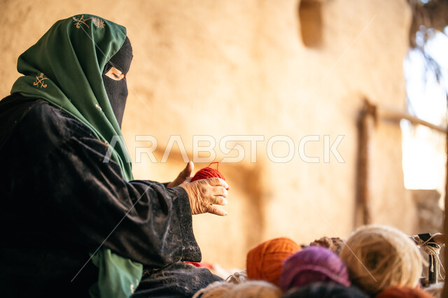 ذكرى تأسيس الدولة السعودية الأولى 1727م, حرفة يدوية تاريخيه, يوم بدينا 22 فبراير، الشغف في ممارسة مهنة الغزل و النسج, المحافظة على التراث الشعبي السعودي, صورة من الجانب لامرأة عربيه خليجية سعودية ترتدي البرقع و العباءه السوداء تجلس في بيت من الطين تقوم بلف خيوط الصوف