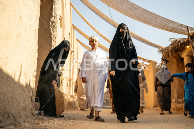 ذكرى تأسيس الدولة السعودية الأولى1727 م، يوم بدينا22 فبراير، مجموعة من الأشخاص في سوق شعبي قديم، مظلة قماشية ممتدة بين البيوت، رجل يرتدي البشت والغترة يمسك بيد ابنته ويتجولان في الحي التاريخي، امرأة عربية خليجية سعودية ترتدي العباءة والنقاب تكنس بمكنسة من القش